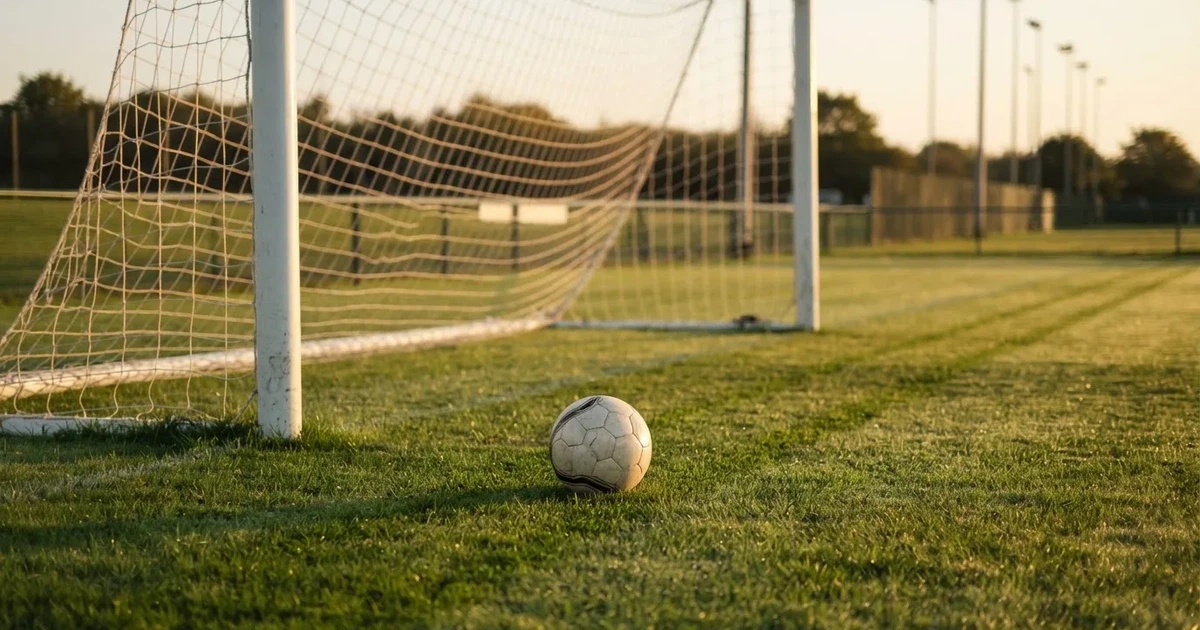 Ein Fußball liegt vor einem leeren Tor auf dem Rasen, das Netz hängt unbewegt, im Hintergrund ein verschwommener Zaun in warmem Abendlicht.