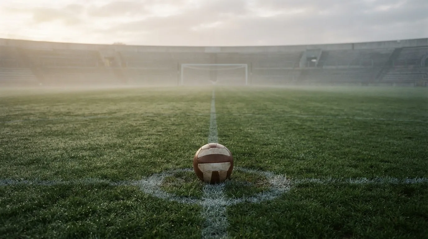 Ein Fußball liegt auf dem Anstoßpunkt eines leeren Rasenplatzes im Morgendunst, im Hintergrund eine ferne Tribüne.