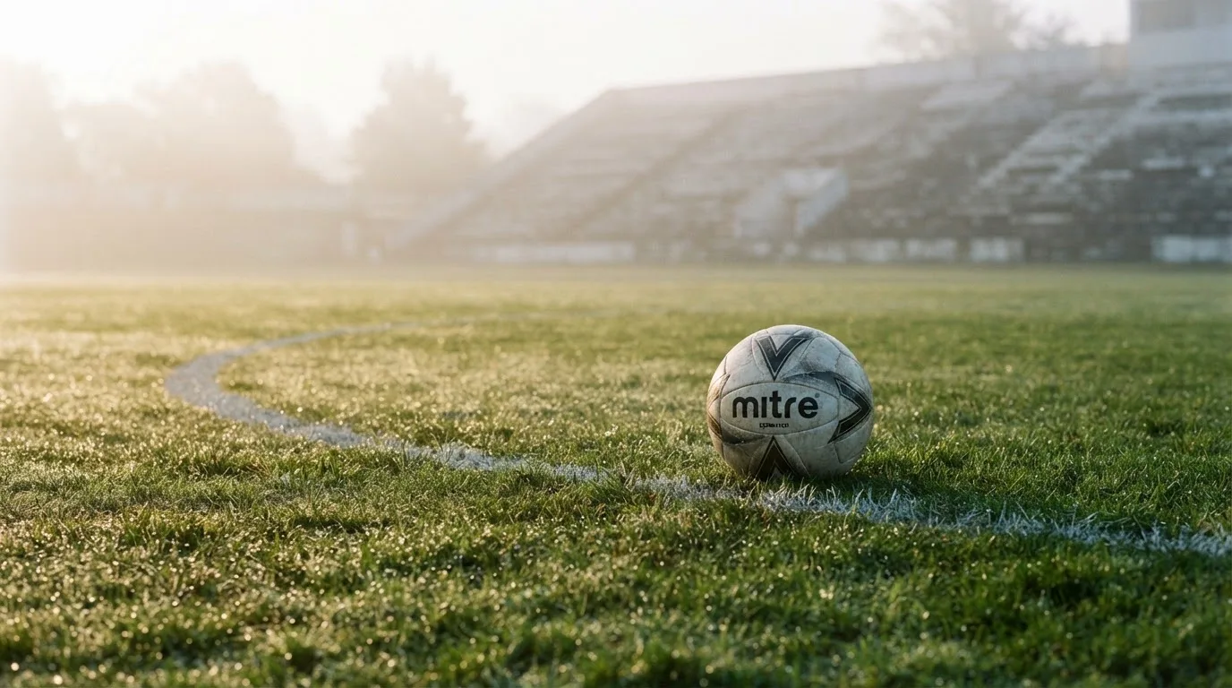 Ein Fußball liegt auf einem Rasenplatz, im Hintergrund eine ferne Tribüne im Morgendunst, Tau glänzt im Gras.