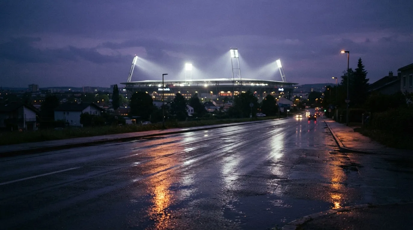 Ein Stadion unter Flutlicht aus großer Entfernung gesehen, davor eine regennasse Straße in der Dämmerung.