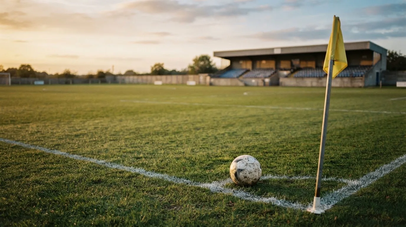 Ein Fußball liegt auf dem Rasen neben der Eckfahne eines leeren Platzes, dahinter eine Tribüne im Abendlicht.