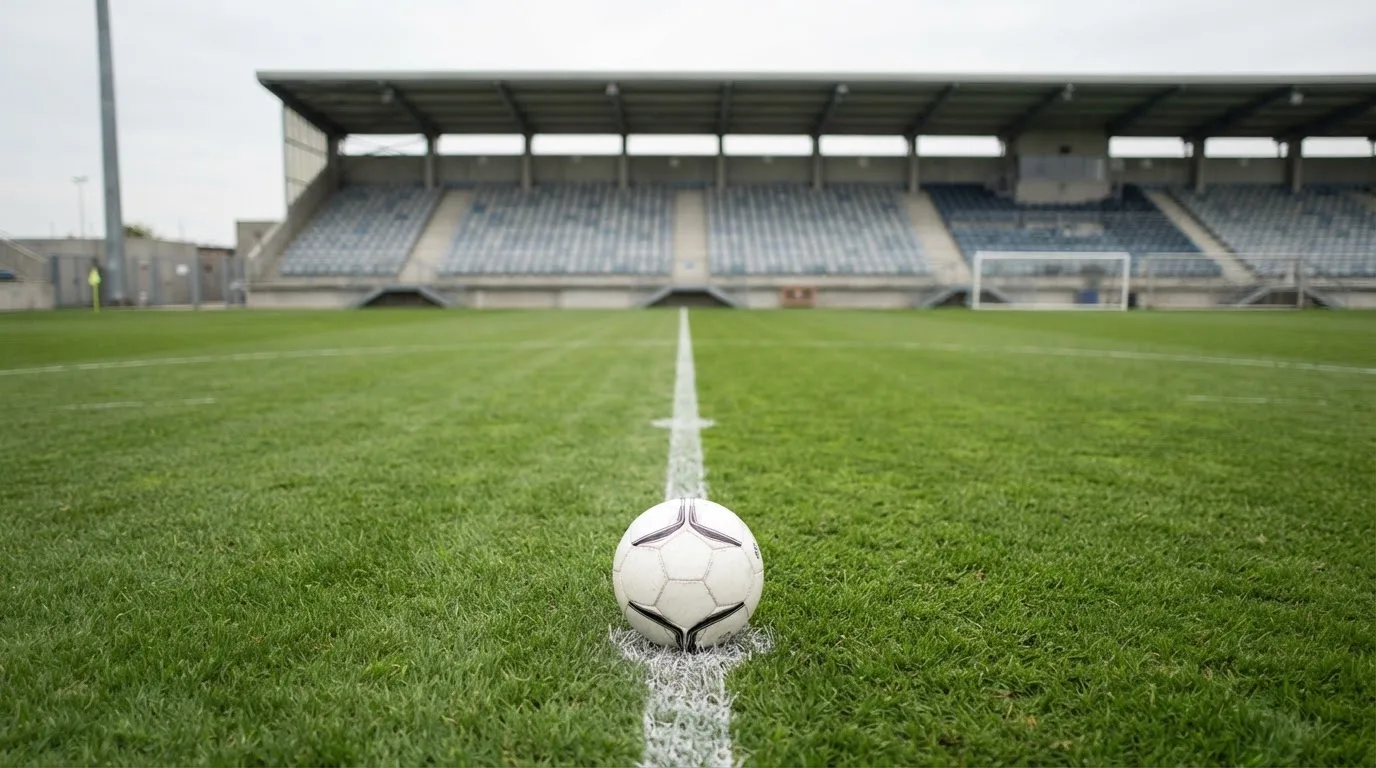 Ein Fußball liegt auf dem Anstoßpunkt eines leeren Rasenplatzes, dahinter eine leere Tribüne in weichem Licht.