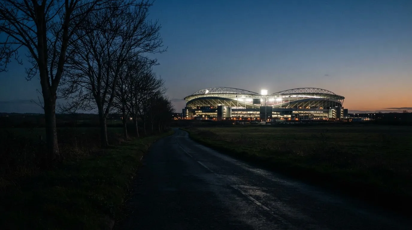 Ein großes Stadion unter Flutlicht aus weiter Entfernung gesehen, davor eine dunkle Landstraße und ein paar Bäume.