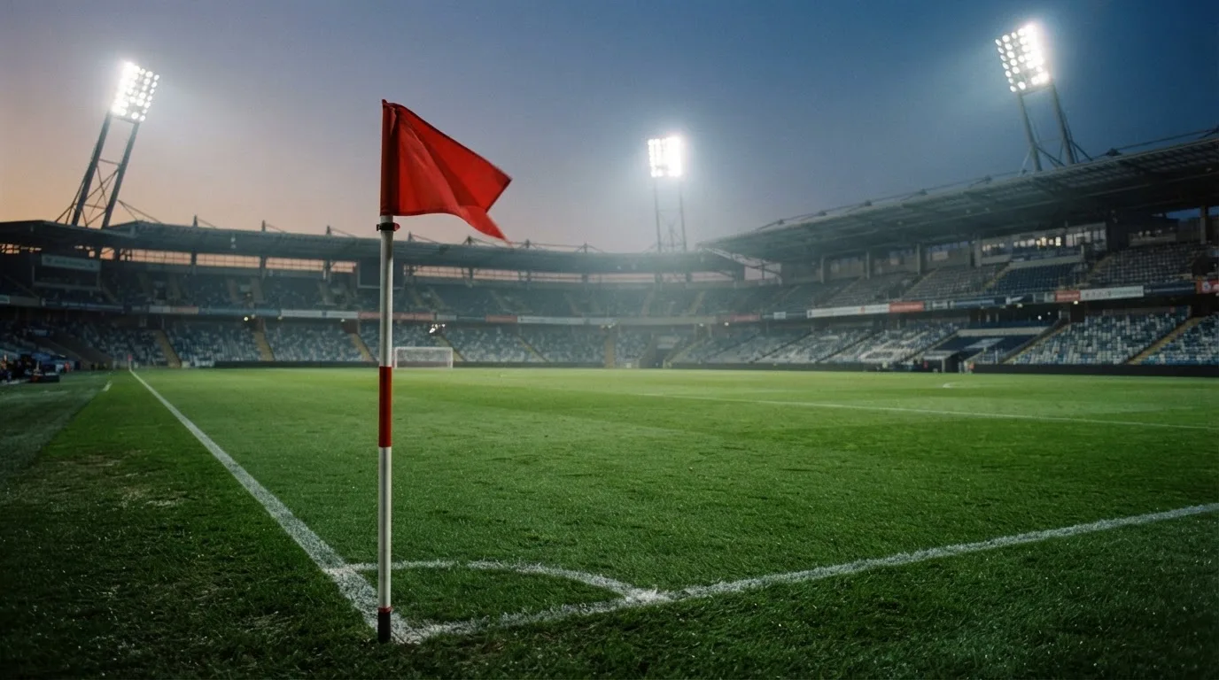 Eine Eckfahne am Rand eines Rasenplatzes, dahinter ein leeres Stadion unter Flutlicht aus der Ferne gesehen.