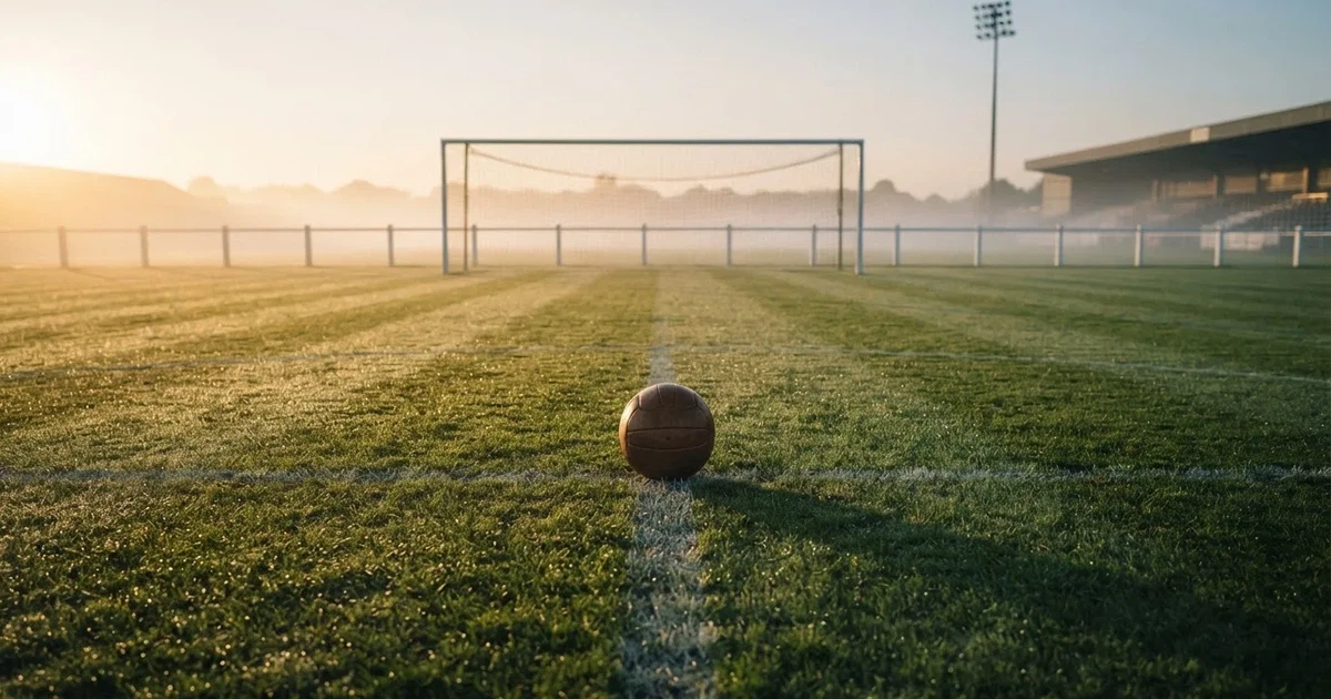 Ein Fußball liegt auf dem Anstoßpunkt eines leeren Rasenplatzes in der Morgendämmerung, Tau glitzert auf dem Gras.