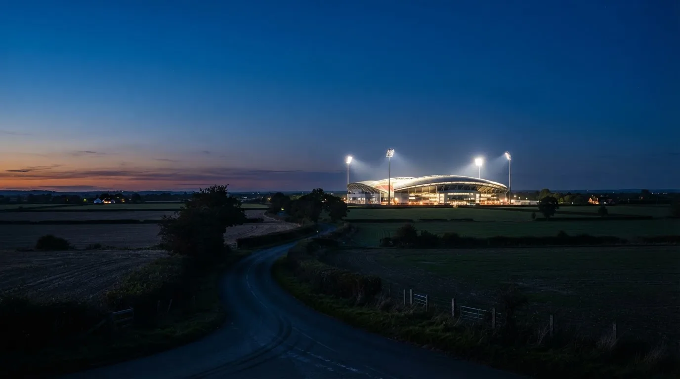Ein Stadion unter Flutlicht aus der Ferne gesehen, davor eine dunkle Landschaft mit Feldern und einer Landstraße.