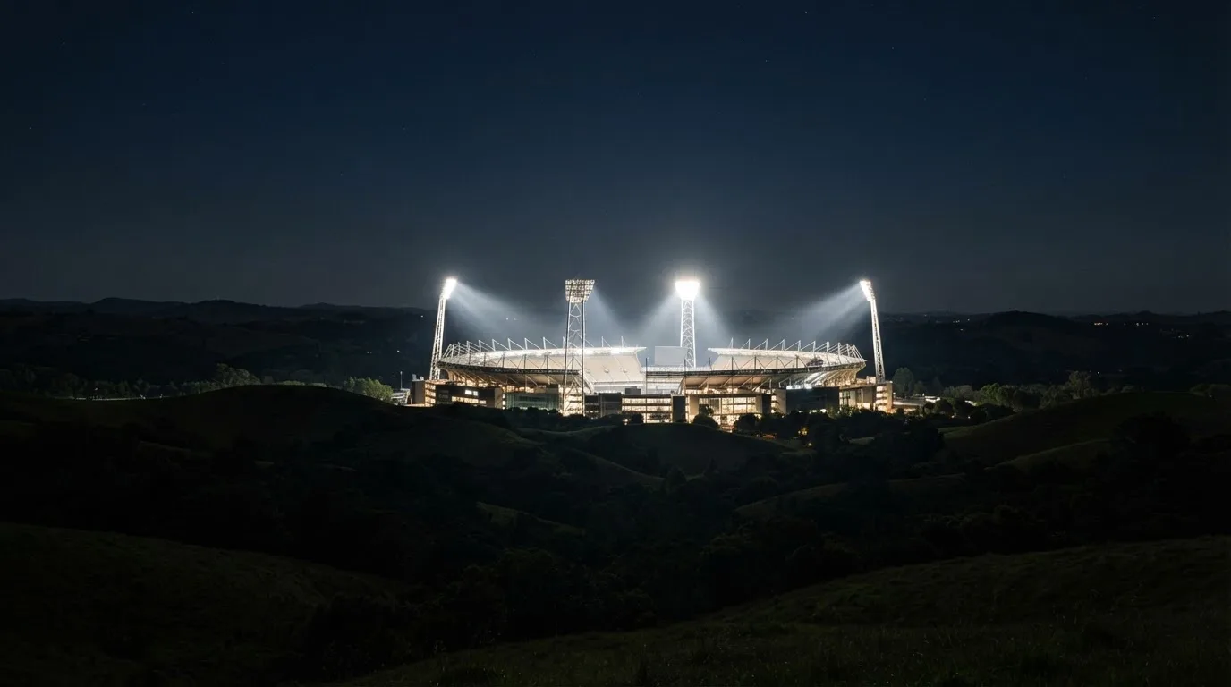 Ein Stadion unter Flutlicht aus großer Entfernung gesehen, darüber ein klarer Nachthimmel und davor dunkle Hügel.