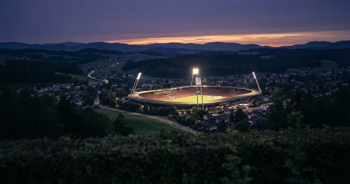 Ein Flutlichtstadion bei Dämmerung aus weiter Ferne gesehen, die Lichtmasten leuchten über Dächern, Bäumen und einer ruhigen Landschaft.