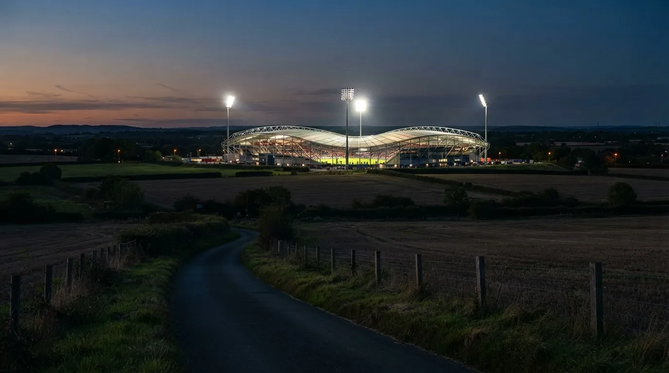 Ein Stadion unter Flutlicht aus der Ferne gesehen, davor eine dunkle Landschaft mit Feldern und einer Landstraße.