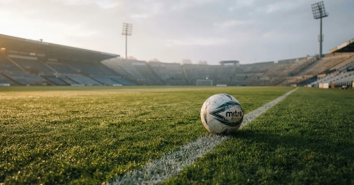 Ein Fußball liegt auf einem Rasenplatz im Morgenlicht, die Kreidelinie verläuft scharf durchs Bild, dahinter eine leere Tribüne.