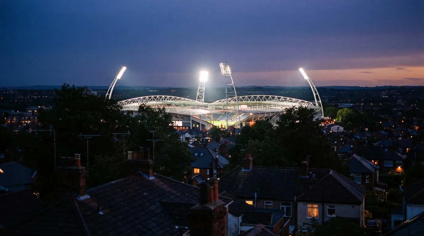 Ein Flutlichtstadion bei Dämmerung aus weiter Ferne gesehen, die Lichtmasten leuchten über Dächern und Bäumen.