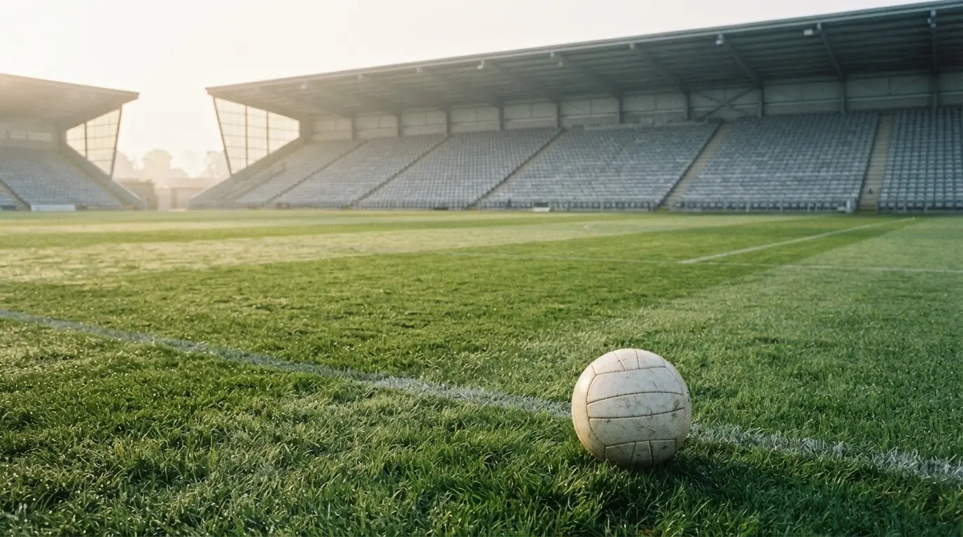 Ein Fußball liegt auf einem Rasenplatz im Morgenlicht, dahinter eine ruhige Tribüne ohne Zuschauer.